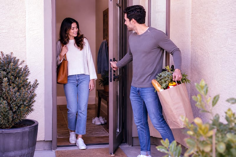A man shows love through actions by carrying groceries and holding the door open for his partner at the entrance of a home, while she smiles softly and notices his thoughtful, protective behavior in warm afternoon light.
